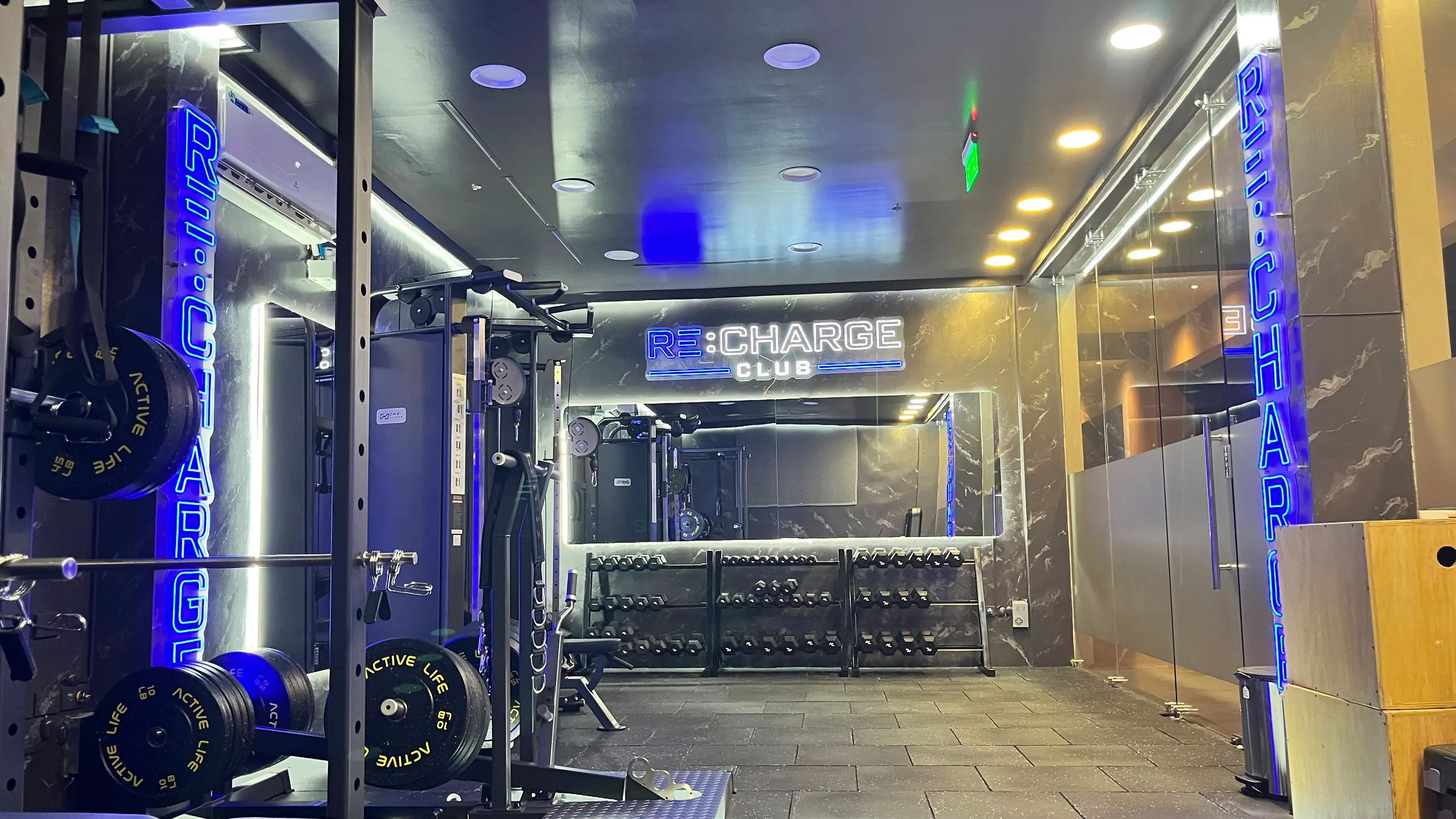 Piso de entrenamiento de la sede Ciudad Celeste: rack con discos, mancuernas y el letrero neón RE:CHARGE CLUB al fondo